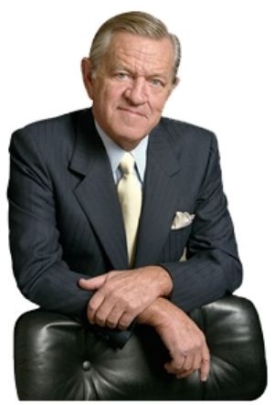 Image of Bill Daniels with a white background. He is leaning on black leather wingback placed in front of him. Daniels is an older white male wearing a dark great suit with white collar shirt and white tie.
